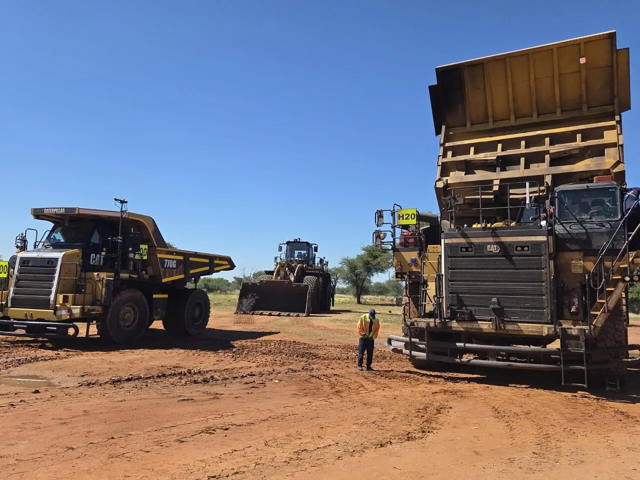 A person on foot standing between haul trucks and a loader at a mine site